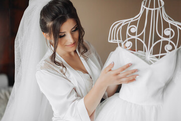 Brunette bride in a robe, posing for the camera, looking at her wedding dress. Elegant hairstyle. Nice makeup. Voluminous veil. Morning of the bride