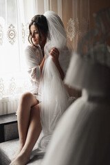 Fototapeta premium Portrait of the bride. A brunette bride in a dressing gown sits behind a dress mannequin, poses, holds her voluminous white veil and looks down. Gorgeous make-up and hair. Wedding photo.