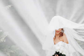 Curly brunette bride in a white dress, covered with a veil, poses for the camera with a bouquet of roses. Portrait of the bride. Beautiful makeup and hair. Wedding in nature