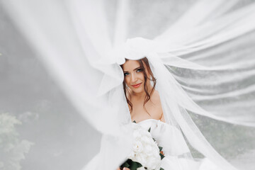 Curly brunette bride in a white dress, covered with a veil, poses for the camera with a bouquet of roses. Portrait of the bride. Beautiful makeup and hair. Wedding in nature