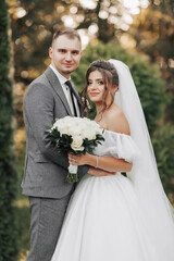 Portrait of the bride and groom in nature. Cropped photo. The bride and groom pose, hugging and smiling, against the background of conifers. The bride in a long dress with a bouquet of roses.