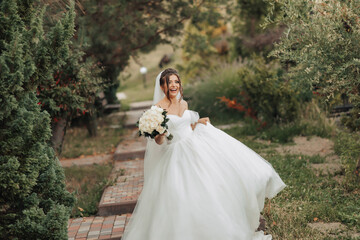 Fototapeta na wymiar Wedding photo. The bride in a voluminous white dress and a long veil, smiling, twirling with a bouquet of white roses, holding her dress. Portrait of the bride. Beautiful makeup and hair.