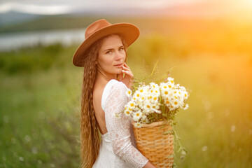 A middle-aged woman in a white dress and brown hat stands on a green field and holds a basket in her hands with a large bouquet of daisies. In the background there are mountains and a lake.