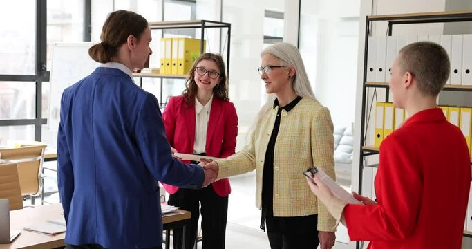 Elegant mature boss-woman in glasses shakes hands with young man to greet near colleagues in office. Employer and employee make friendly handshake slow motion