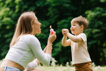 Young Caucasian mother and her happy little son are having fun playing with soap bubbles in the park