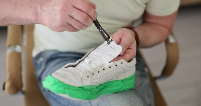 Man paints sneaker with sole taped green using thin brush and white dye. Male person sits on chair and renovates shoes at home slow motion