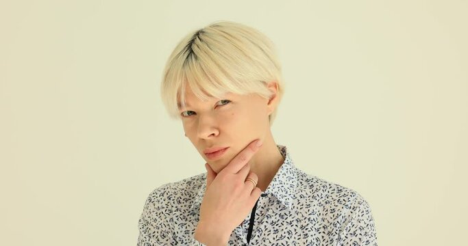 Blonde woman with short stylish hairdo suspiciously looks and squints. Lady in patterned blouse puts hand up to face while standing near wall