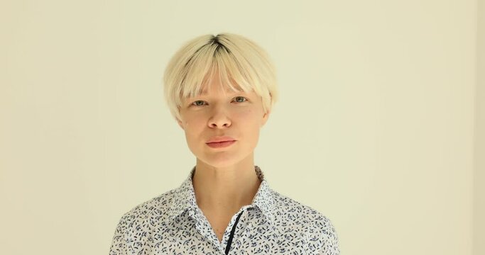 Stylish young woman in patterned office blouse winks and looks skeptically. Stylish woman with short blonde hairdo expresses mistrust against beige wall