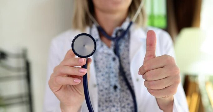 Pleased doctor holds stethoscope in hand showing thumb-up. Woman approves serviceability of medical equipment standing in clinic office slow motion