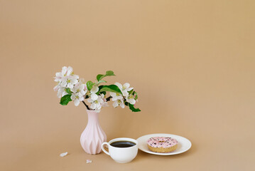 Beautiful white apple tree flowers in a white vase, donuts and a cup of tea on a beige background with a copy space