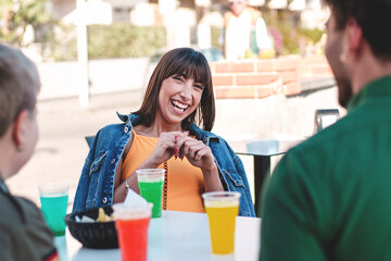 Smiling Woman at Seaside Bar - Young brunette woman laughing, wearing an orange shirt and denim jacket, with her friends at a seaside bar terrace.
