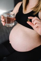 Fototapeta premium Pregnant woman take vitamins daily Omega 3 and glass water. Female hands hold supplements or vitamin. Girl sitting in bed at home. Pregnancy, treatment, medicine healthcare concepts. Photo of pills.