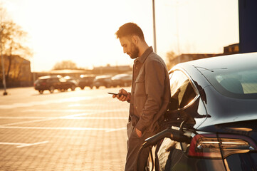 On the parking lot. Charging the automobile. Man is standing near his electric car outdoors