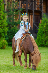 A little girl in the Ukrainian national costume rides a pony on the lawn