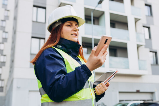 Architect woman in white hardhat and safety vest with digital tablet using smartphone outdoors. Female engineer. Logistics and construction worker near modern building. Engineering application