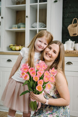 Daughter congratulates mother and gives bouquet of tulip flowers in kitchen at home. Mother's day concept. Mom hug child girl smiling and surprised. Greeting card. International Women's Day. Closeup