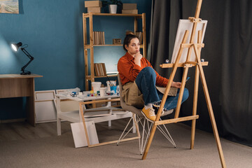 young brazilian woman artist in home studio sits on a chair near an easel and thinks over the idea of a new painting. . concept of creativity and hobby