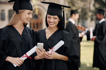 Two happy young women in mortarboards holding diplomas and looking at smart phone