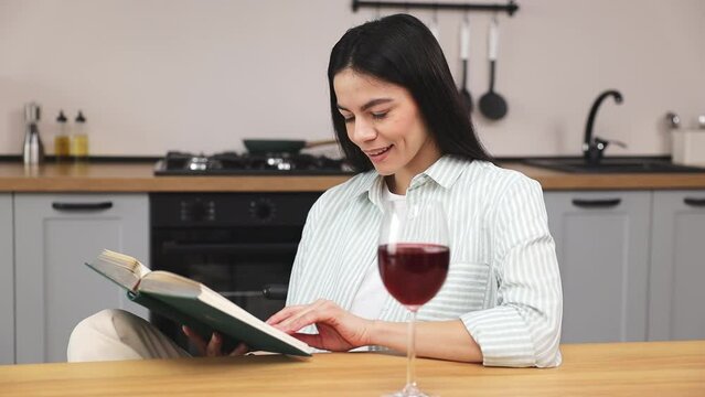 Young calm happy brunette housewife woman 30s wear casual clothes shirt read book novel hold glass drink red wine relax rest sit at table in light kitchen at home alone. Lifestyle cooking food concept