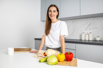 Smiling young woman standing near table with fresh vegetables in kitchen