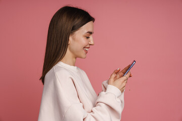 Fototapeta premium Profile of smiling woman using mobile phone while standing isolated over pink background