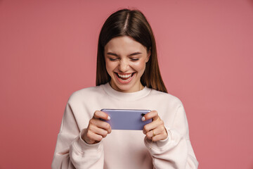 Excited woman playing online game on smartphone isolated over pink background