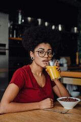 African woman drinking orange juice and eating cereal while sitting in kitchen
