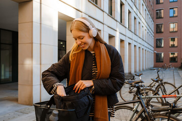 Young woman in headphones putting something in her backpack while standing with bicycle outdoors
