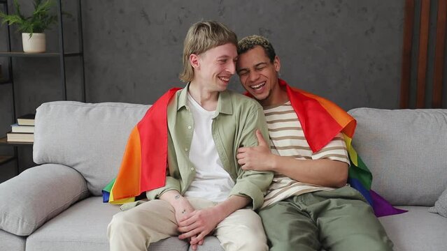 Young happy couple two gay men wear casual clothes covered striped flag sits on sofa couch stay at home flat relax spend free spare time in living room indoor. Pride day june month love lgbtq concept