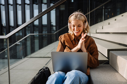 Smiling woman in headphones making video call via laptop and gesturing while sitting on stairs
