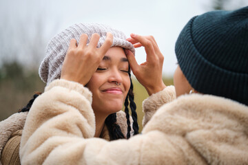 Dominican lesbian woman putting a woolen hat on her girlfriend head at street in winter.
