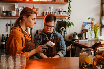 Woman teaching man employee with down syndrome how to use payment terminal in coffee shop