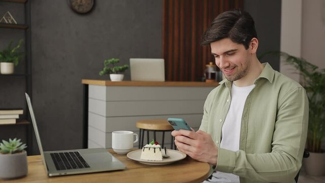 Young man 20s wear casual clothes sits alone at table in coffee shop cafe work on laptop pc chatting typing message on cell phone relax rest in restaurant during free time indoor Mobile office concept