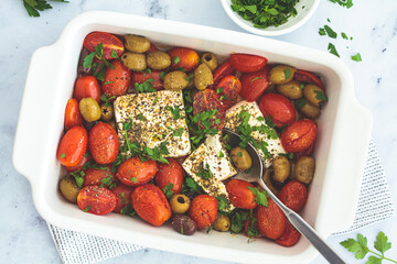 Baked feta cheese with cherry tomatoes and olives on a white table.