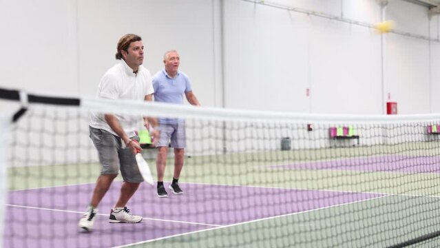 Portrait of sporty adult man playing doubles pickleball with experienced aged partner on indoor court, ready to hit ball. Sport and active lifestyle concept Generative AI