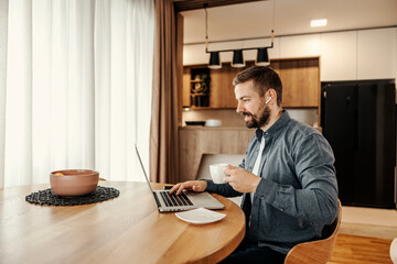 A happy entrepreneur is enjoying his coffee while working from home on a laptop.