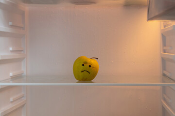 A sad face is drawn on a green apple that stands in the refrigerator on an empty shelf, hunger and...