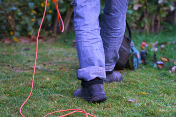 A man in jeans with a lawn mower mows a lawn strewn with fallen autumn leaves. Photo from below, only legs in the frame