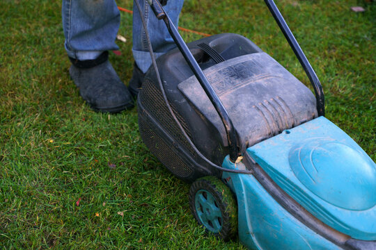 A man in jeans with a lawn mower mows a lawn strewn with fallen autumn leaves. Photo from below, only legs in the frame
