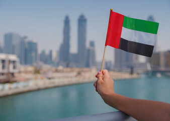 Close up of a female hand holding tiny flag of UAE against Dubai skyline. Photo of UAE national day...