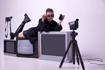 Attractive young girl in stylish black jacket, boots and sunglases lying on retro tv sets and recording video with camera. Concept of fashion, 80s, 90s style, retro and vintage, gadgets, business