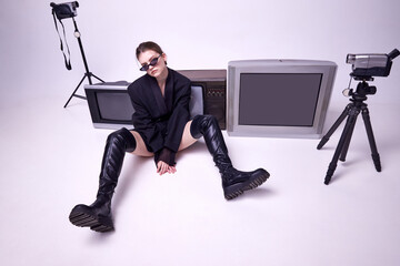 Young girl in stylish black jacket, leather boots and sunglasses sitting on floor around retro TV...