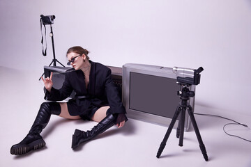 Young girl in stylish black jacket, leather boots and sunglasses sitting on floor around retro TV...