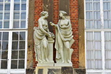 Antique statue of two women on a pedestal at the facade of the building. Brick red wall and tall old windows in a white frame. Monumental art. Germany, Potsdam, Royal Castle, August 2022.