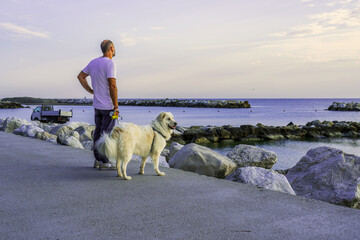 A man with a big white dog on a leash on a beautiful rocky seashore at dawn, walking with a domestic dog, pets, authentic sustainability, sea dawn, unity with nature, copy space - Powered by Adobe