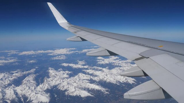 View from passenger cabin window of wing of aircraft flying over Austrian Alps on sunny day against background of blue sky and landscape with snow-capped rocky mountain ranges below. Generative AI