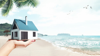 Moving to island. Female hand holding 3D model of small comfortable house against beach, palms and ocean background. Concept of real estate, buying house, mortgage, ownership, business - obrazy, fototapety, plakaty