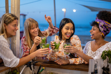 Group of beautiful young women participate in a beach party at night, best female friends toast with cocktails and mojitos, summer holiday concept and carefree