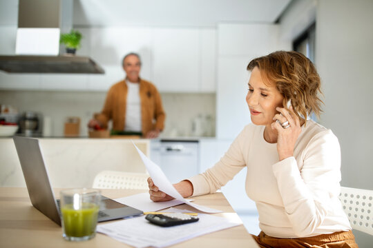 Cheerful senior woman talking on phone and holding papers while her husband preparing dinner at kitchen, selective focus Generative AI