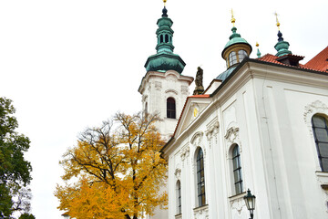 View of the old catholic church with white walls, red tiled roof and green tower, and a tree with yellow leaves. Autumn view, cloudy sky. Prague, Czech Republic, October 2022.